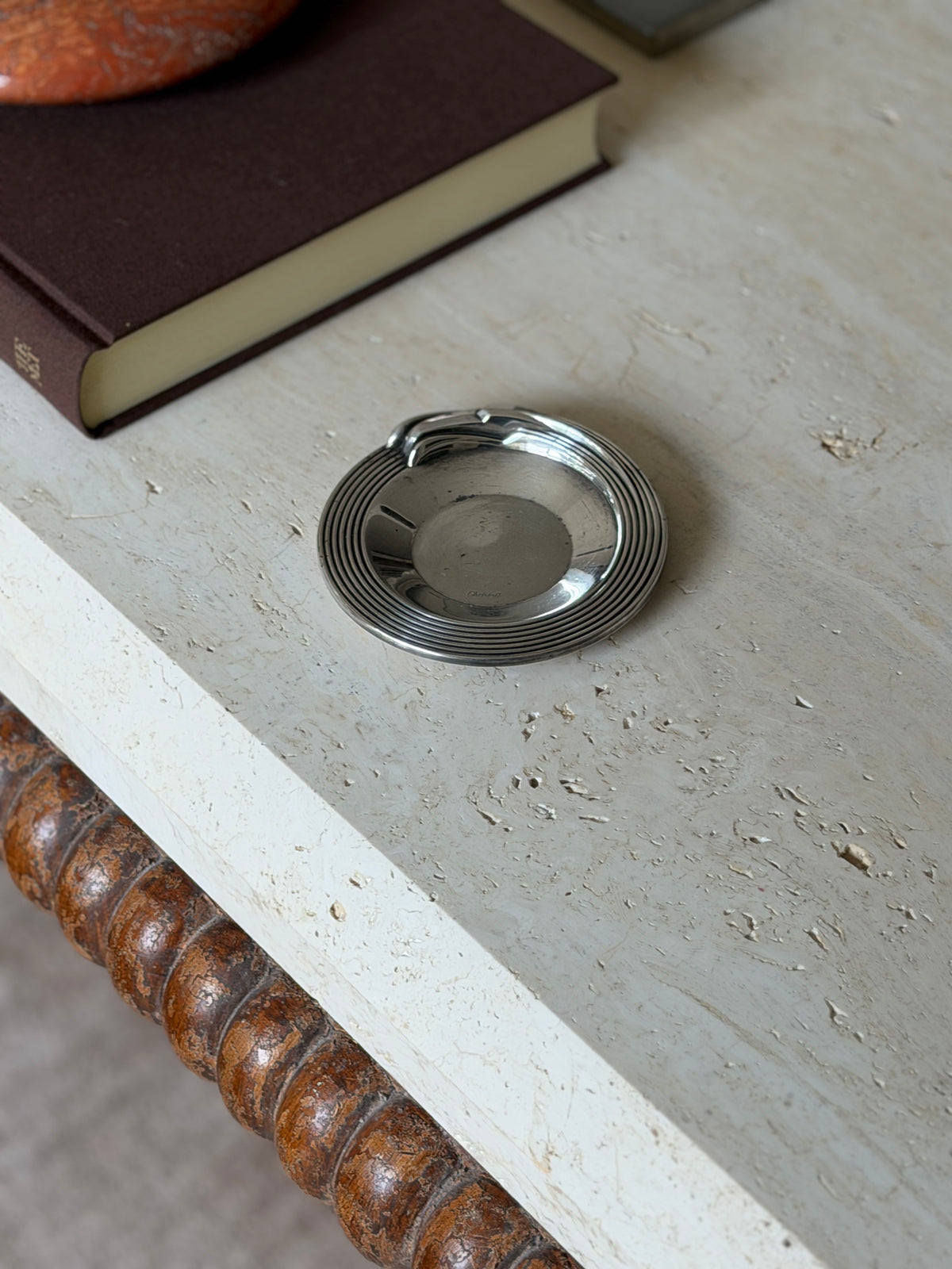 High angle view of the round silver plated Christofle dish on travertine beside a brown book and red stone ashtray, a bobbin turned apron below