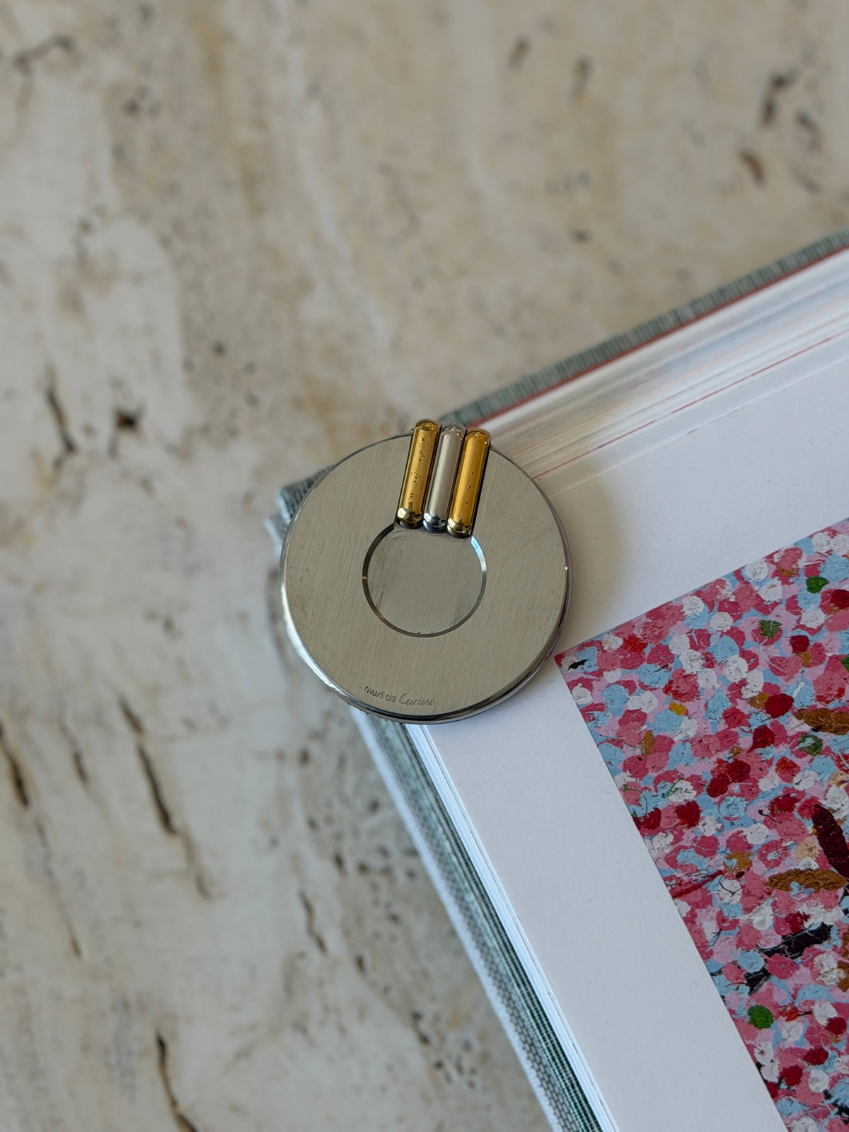 Must de Cartier cigar cutter on the corner of an art book, a brushed steel disc with a round cutting hole and three gold and silver tone bars, signed must de Cartier