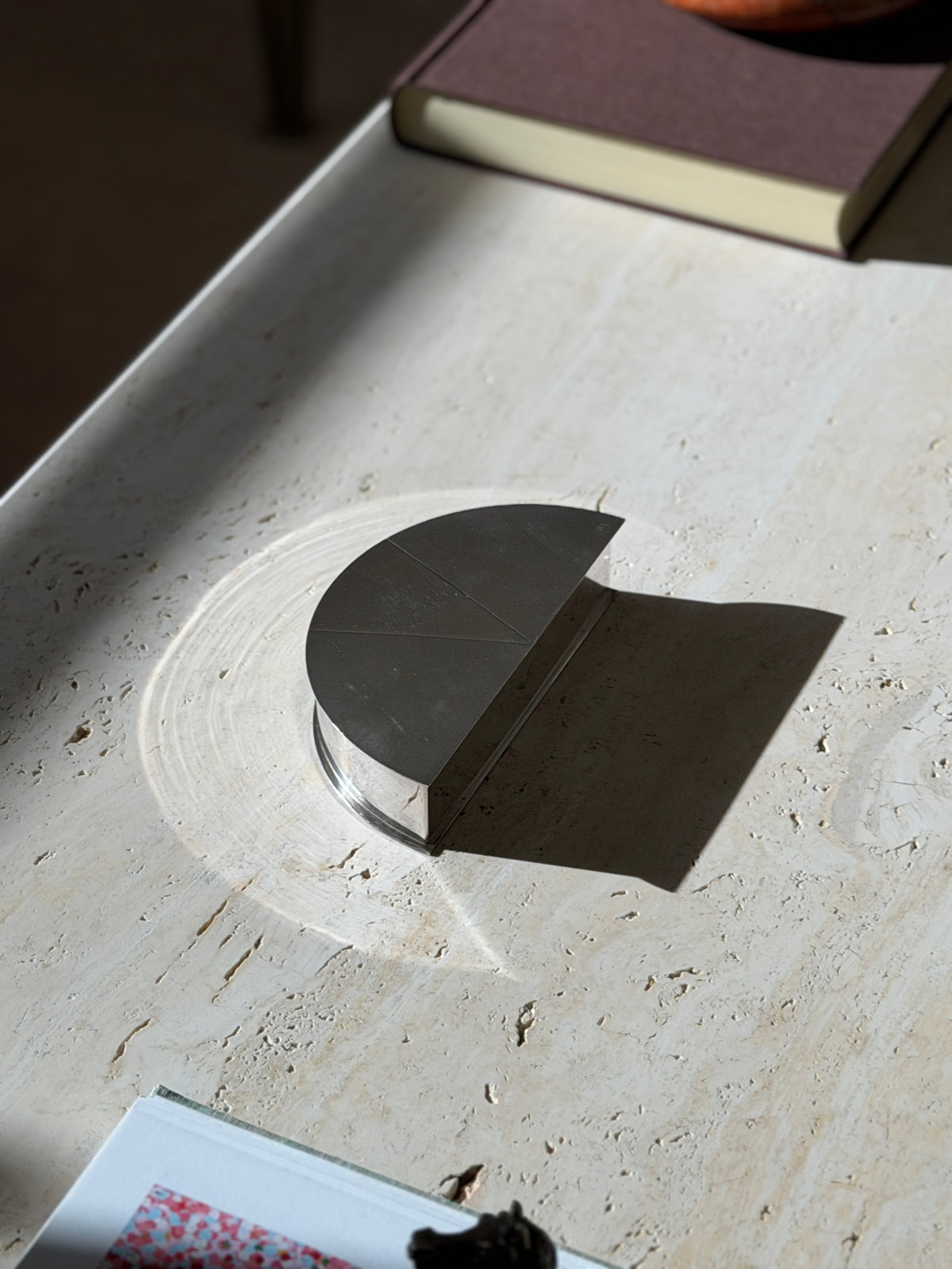Olivetti modernist box on a travertine table casting a crisp shadow, the stepped polished base visible below the textured lid
