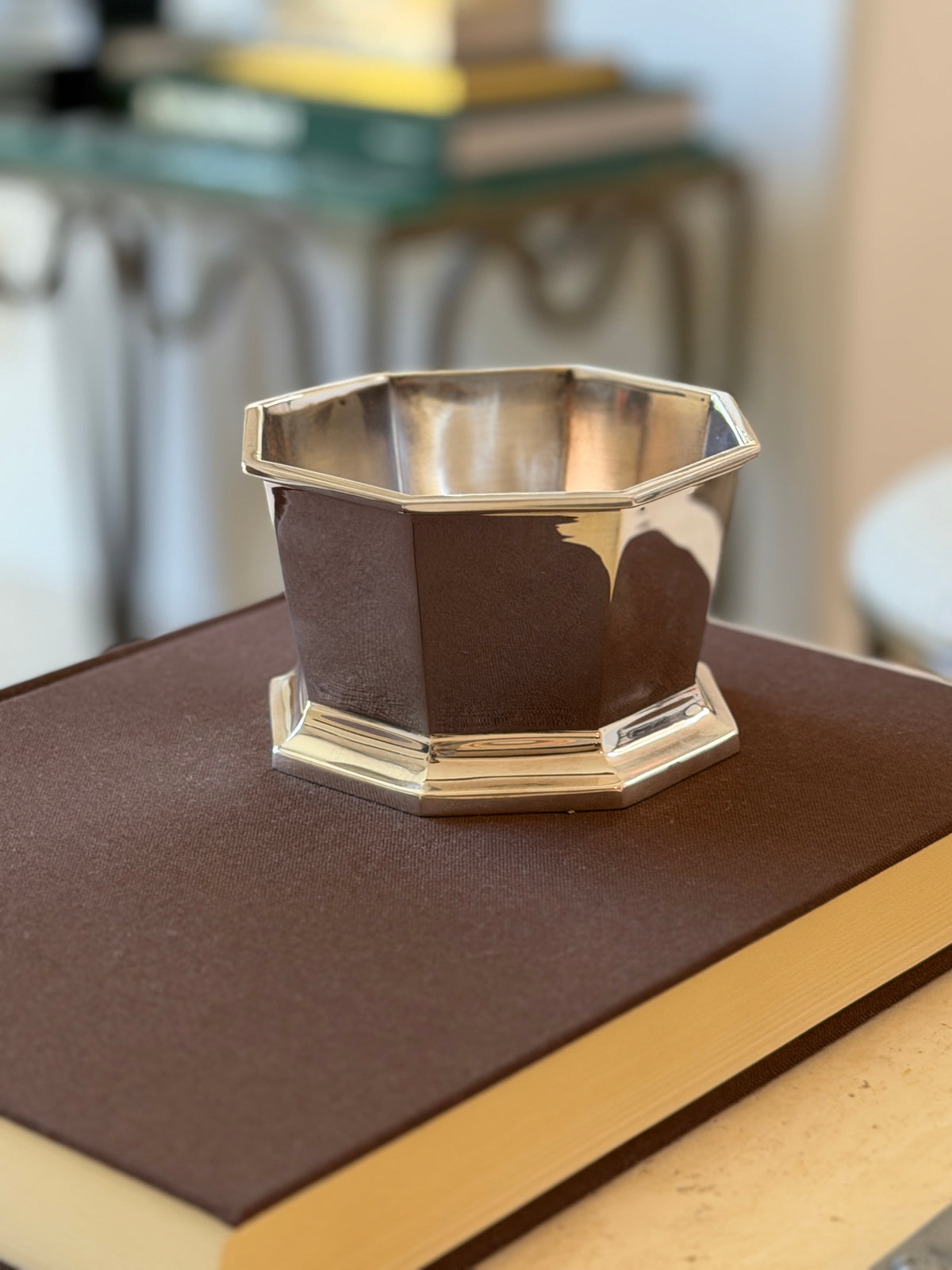 Ralph Lauren octagonal silver bowl on a brown book atop a console, a stack of books and a glass table blurred behind