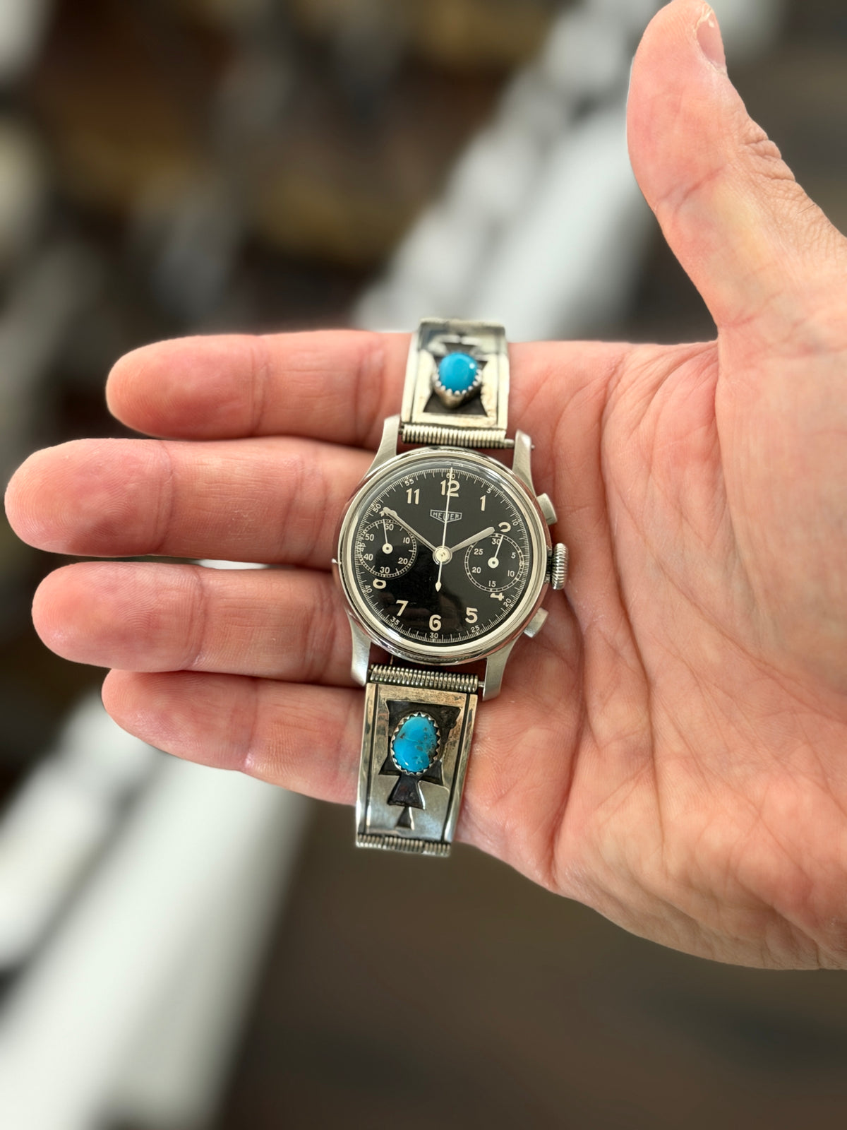 Front view of 1940s Heuer black dial chronograph on stamped sterling silver turquoise bracelet held in palm