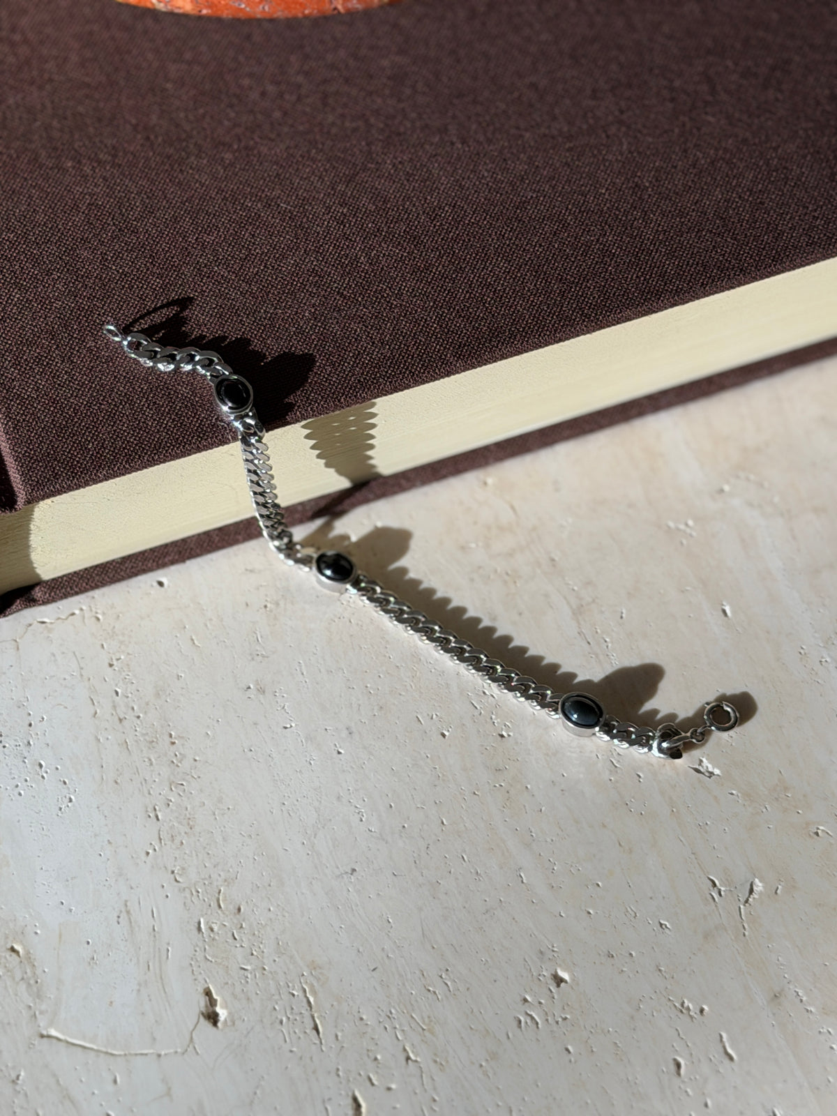 Overhead view of the onyx and silver curb bracelet laid straight on a brown book beneath a red stone ashtray with brass well