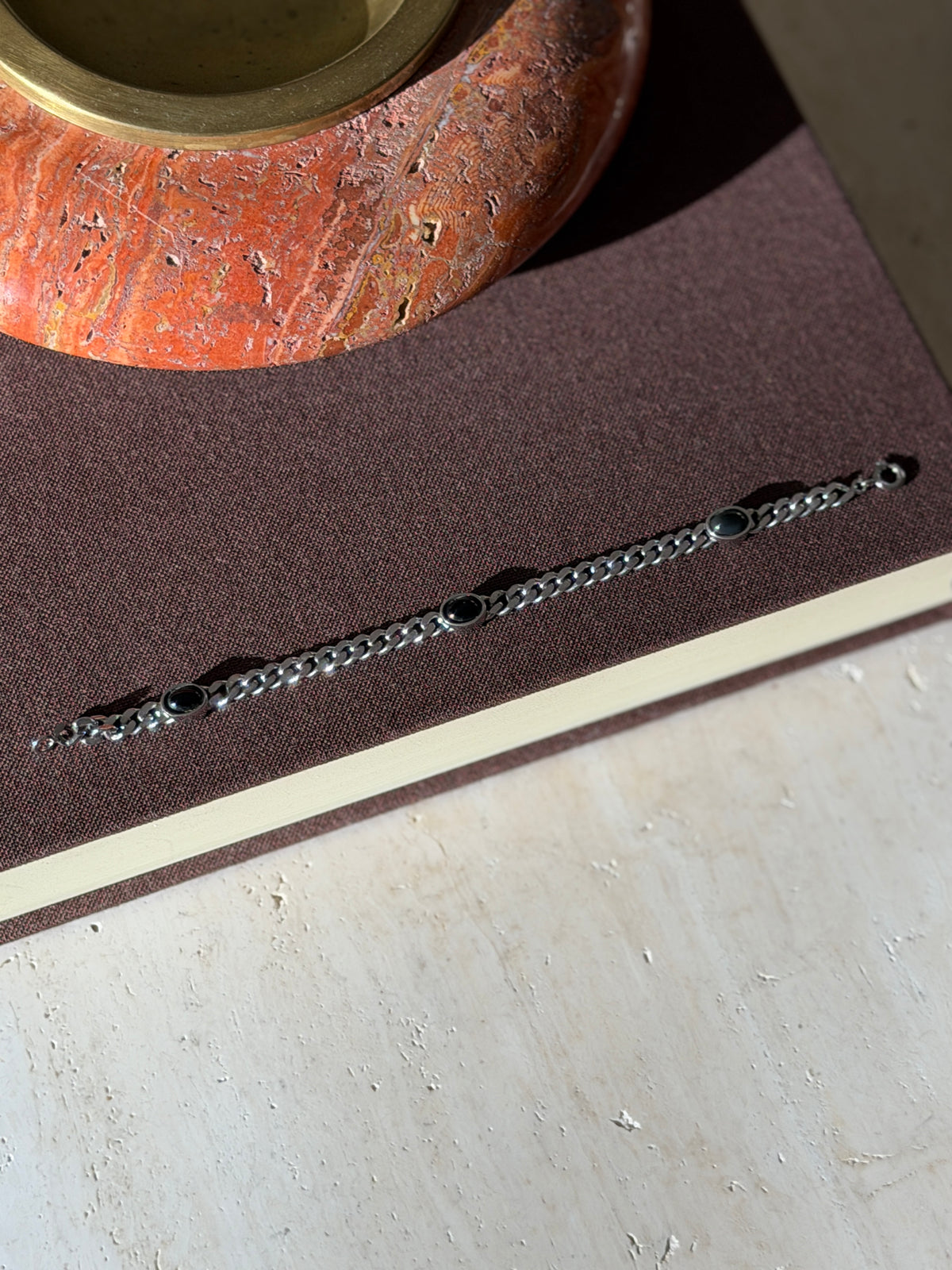 Onyx and sterling curb bracelet laid flat on a brown book in sunlight, three black oval cabochons spaced along the chain below a red stone ashtray