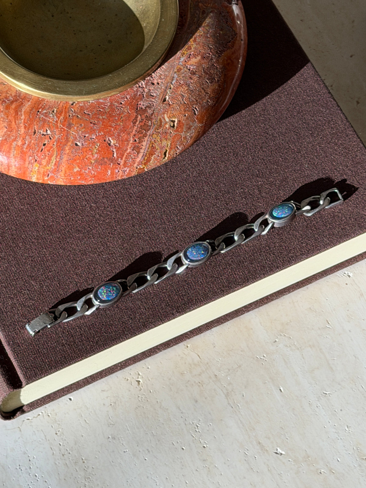 Overhead view of the curb link bracelet with three oval opals on a brown book, a red stone ashtray above in afternoon light