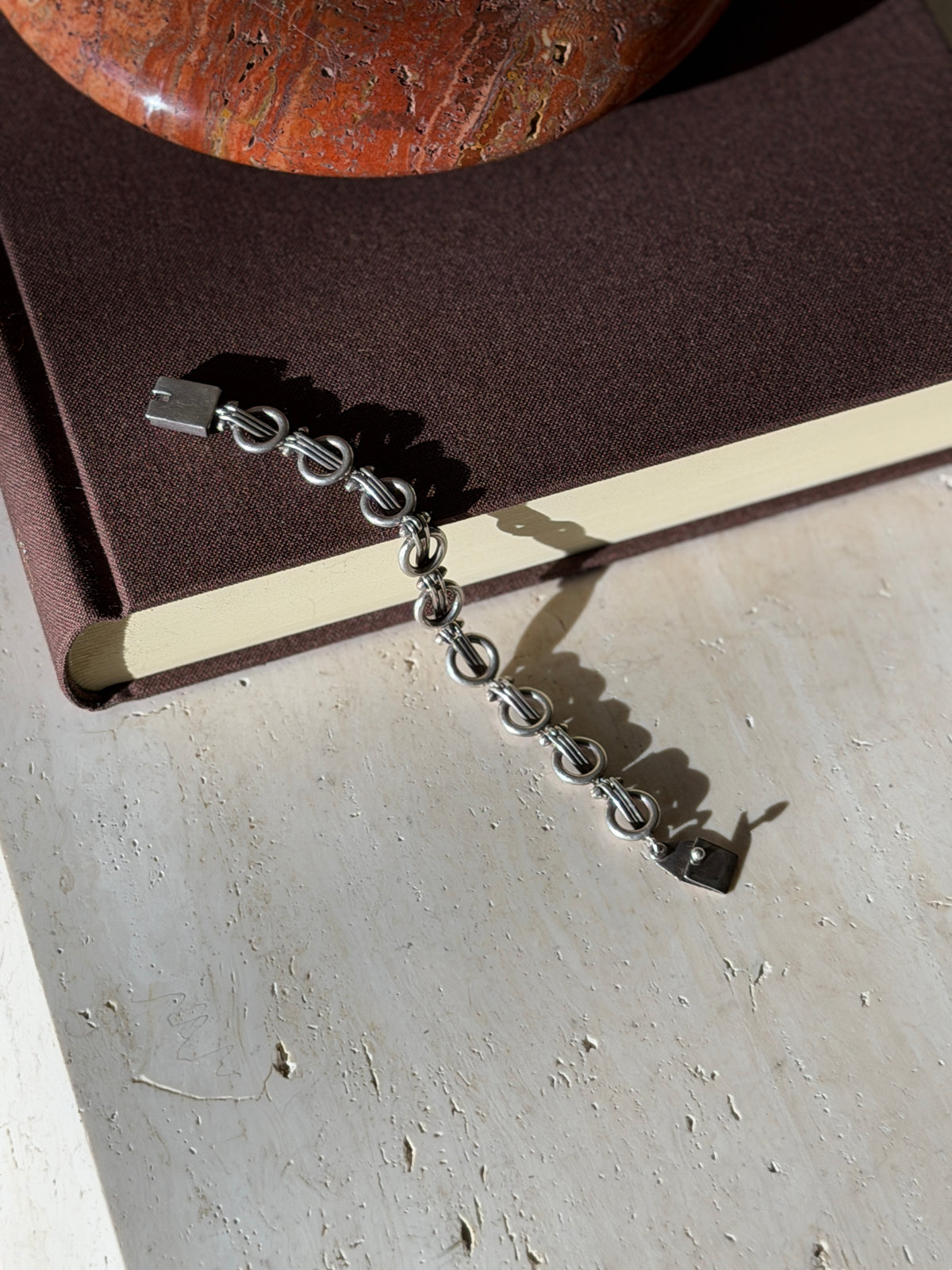 Ring and bar link bracelet draped from a brown book onto travertine, its flat hinged tab clasp at each end