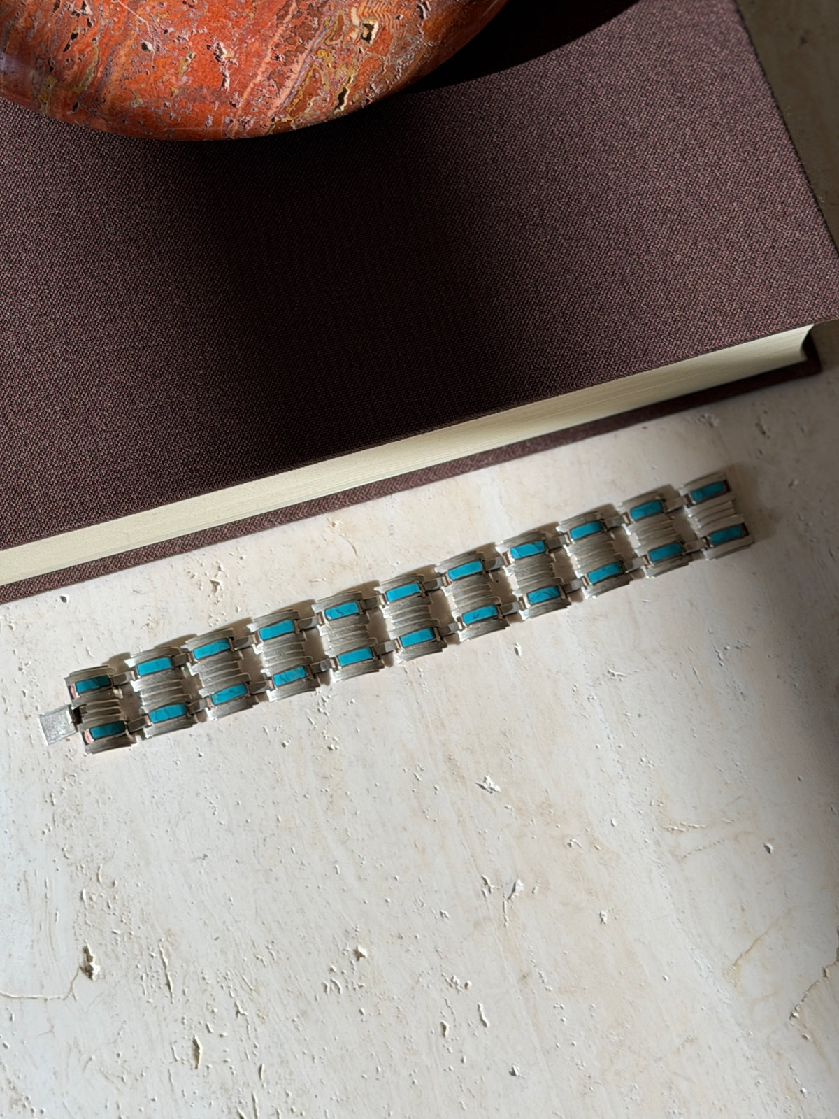 Overhead view of the full Narvaez bracelet laid flat on travertine below a brown book, two rows of turquoise bars along the ribbed links