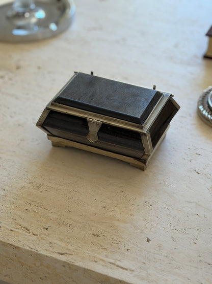 Black stone casket box with a beveled lid, faceted sides, and silver tone mounts, resting on a travertine table beside a glass dish