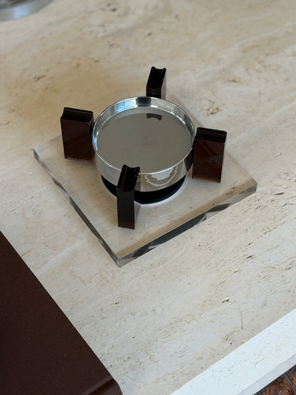 Top-down view of the square base ashtray set diagonally on travertine, the four amber uprights evenly spaced around the reflective chrome bowl