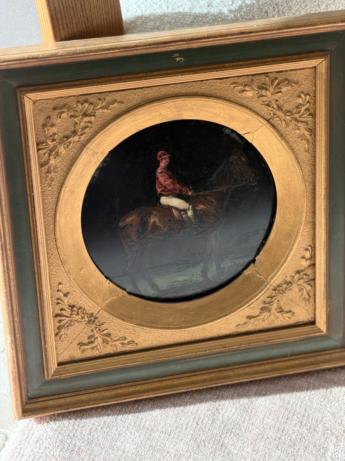 Front view of the jockey painting, the rider in red silks and cream breeches holding the reins against a near black ground, a cracked gilt round mat around the plate