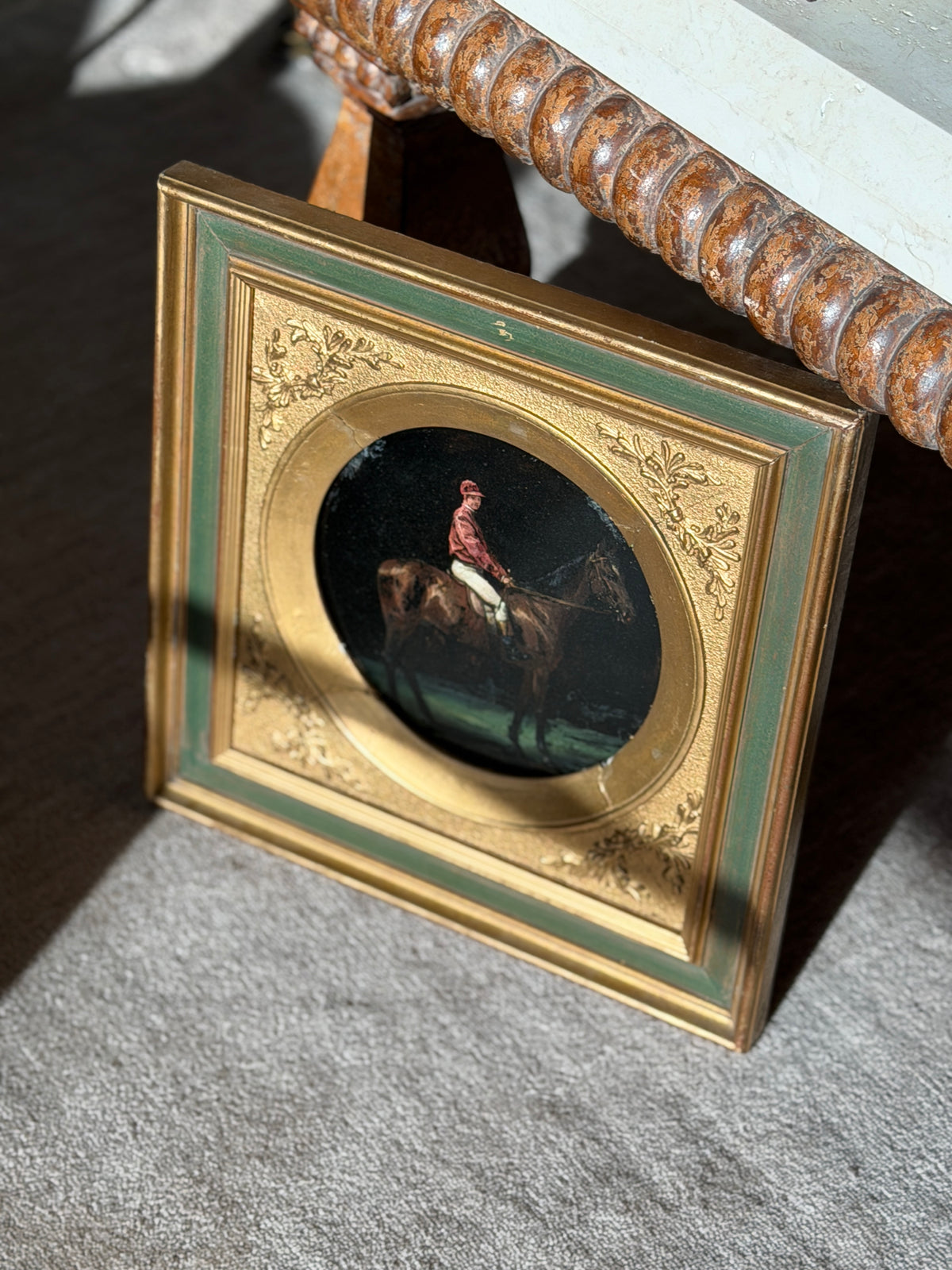 Jockey on horseback painting propped beneath a carved bobbin edge console, sunlight raking across the gilt and sage green frame