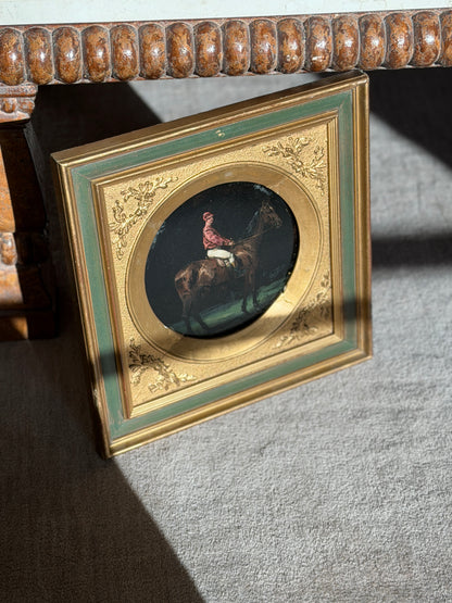 Angled view of the round jockey portrait under a carved wood table, showing the embossed oak leaf corners and patinated gilding