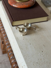 Two small silver dishes with angular geometric handles resting side by side on a stone surface, an open book above them