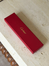 Original red Cartier presentation box with gold script logo on the grained lid, resting on a travertine table above a bobbin turned wood frame
