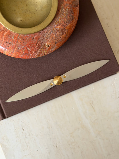 Overhead view of the steel and gold propeller letter opener on a brown linen book cover, the twin tapered blades set on a diagonal