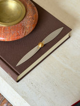 Cartier letter opener shaped like an aircraft propeller, styled on a brown book near a red stone ashtray with the edge of the red box in view
