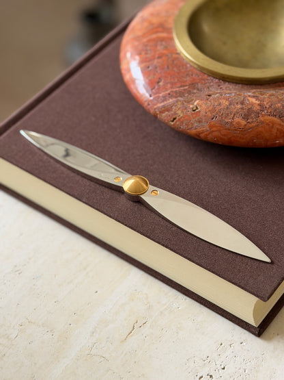 Angled view of the propeller letter opener along the book edge, light running down the polished steel blade toward the gold center hub