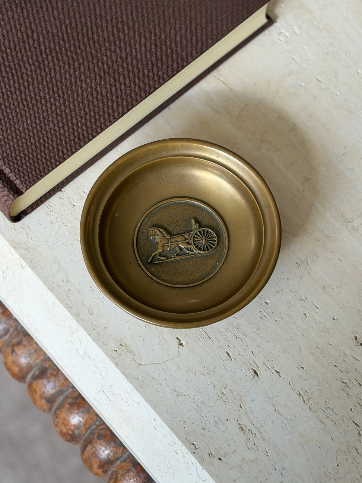 Overhead view of the brass dish with its embossed horse-drawn carriage medallion, resting on a book near the table edge