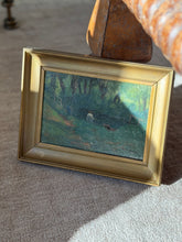 Ceneri Morand woodland pond oil painting in a deep gilt frame leaning against a rust textured chair base on a pale rug, sunlight across the green canopy