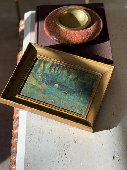Morand landscape painting in a stepped gilt frame on a travertine table, a red stone ashtray with brass well on a brown book behind