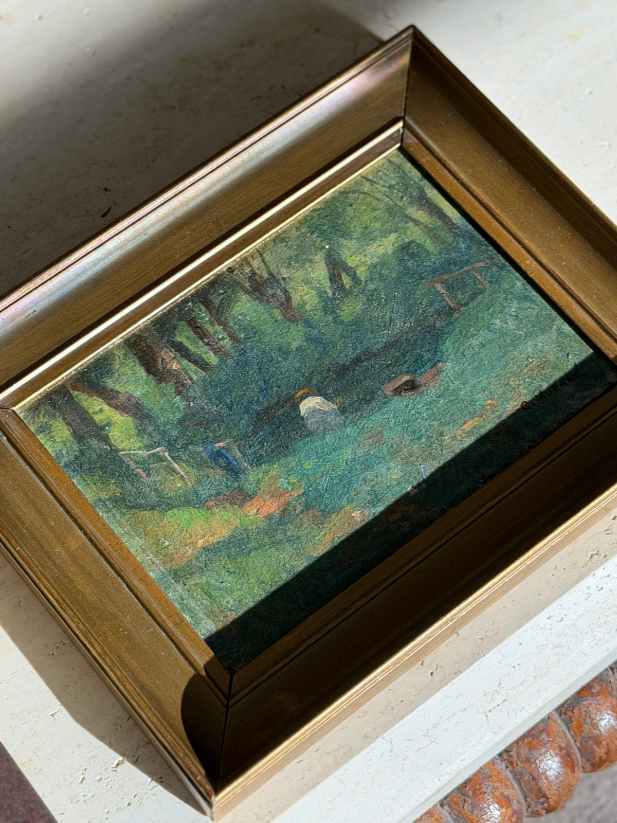 Framed oil painting of a secluded woodland pond laid flat on a pale rug, tall trees and a winding orange earth path, signature at lower right