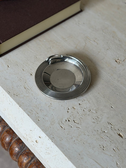 Christofle Art Deco ashtray dish on a travertine table, concentric ribbed rim sweeping into a sculpted rest, Christofle stamped in the polished well