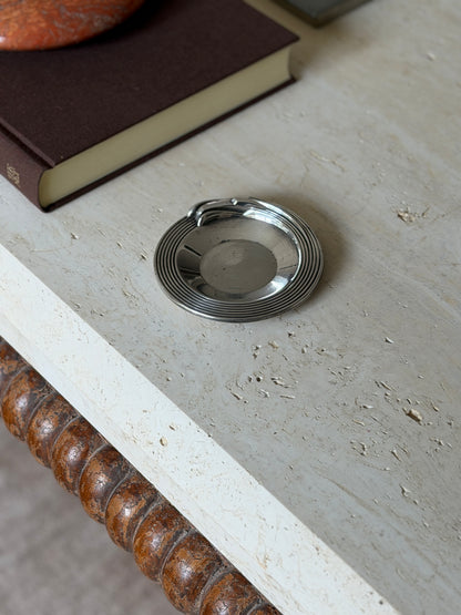 High angle view of the round silver plated Christofle dish on travertine beside a brown book and red stone ashtray, a bobbin turned apron below