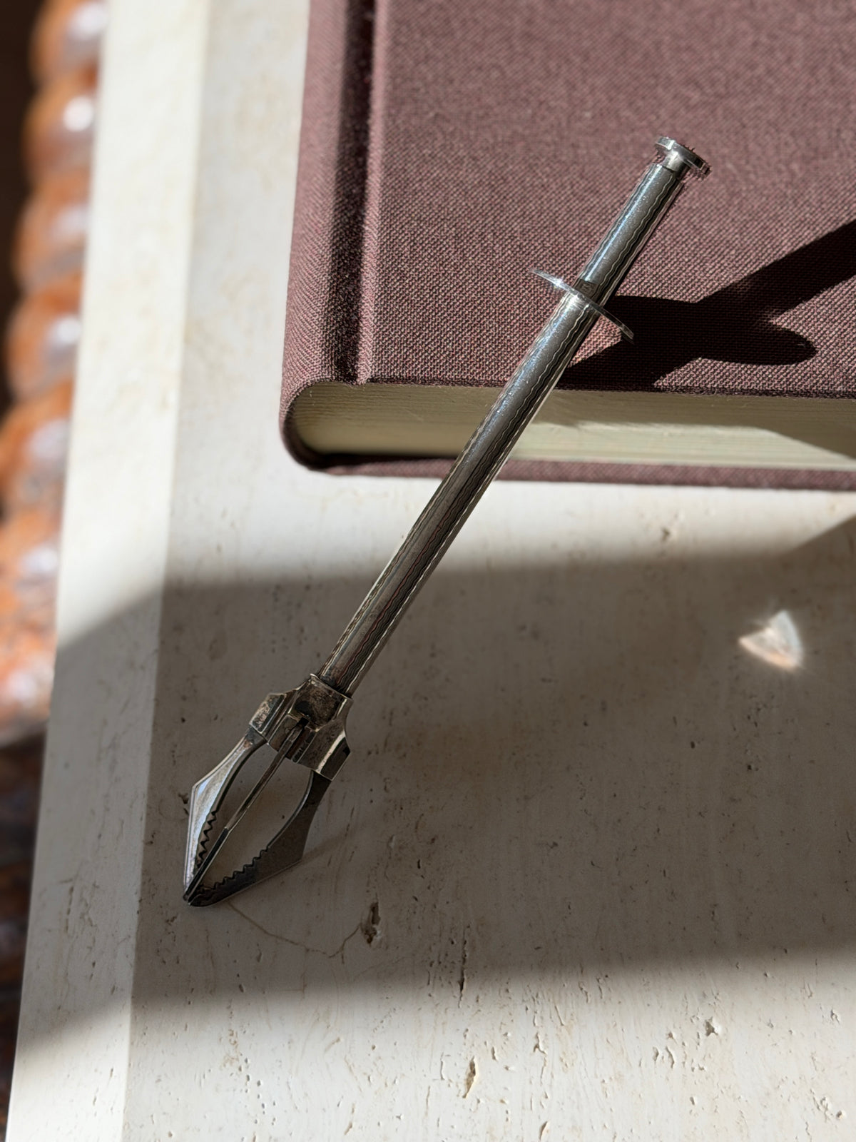 French mechanical tongs propped on a book edge, light catching the engine turned pattern and the open claw grips