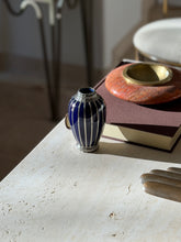 Small blue and silver overlay vase at the edge of a travertine console near a brown book and red stone ashtray, a chair blurred behind