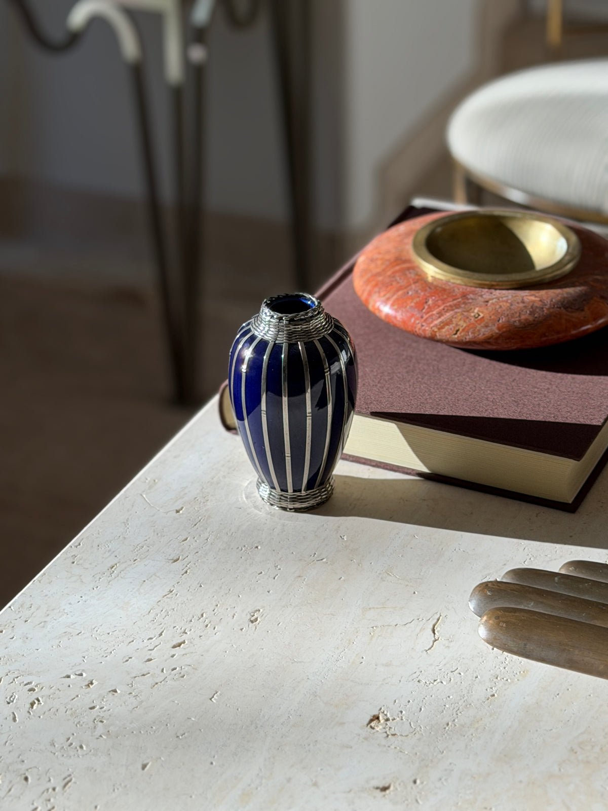 Small blue and silver overlay vase at the edge of a travertine console near a brown book and red stone ashtray, a chair blurred behind