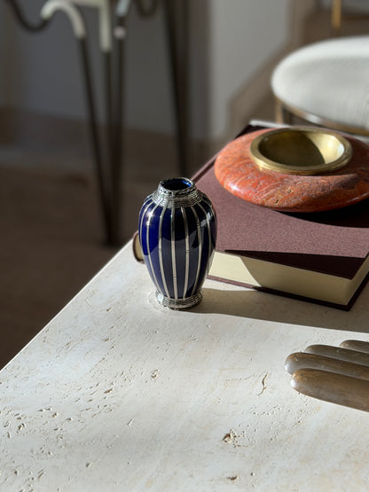Small blue and silver overlay vase at the edge of a travertine console near a brown book and red stone ashtray, a chair blurred behind