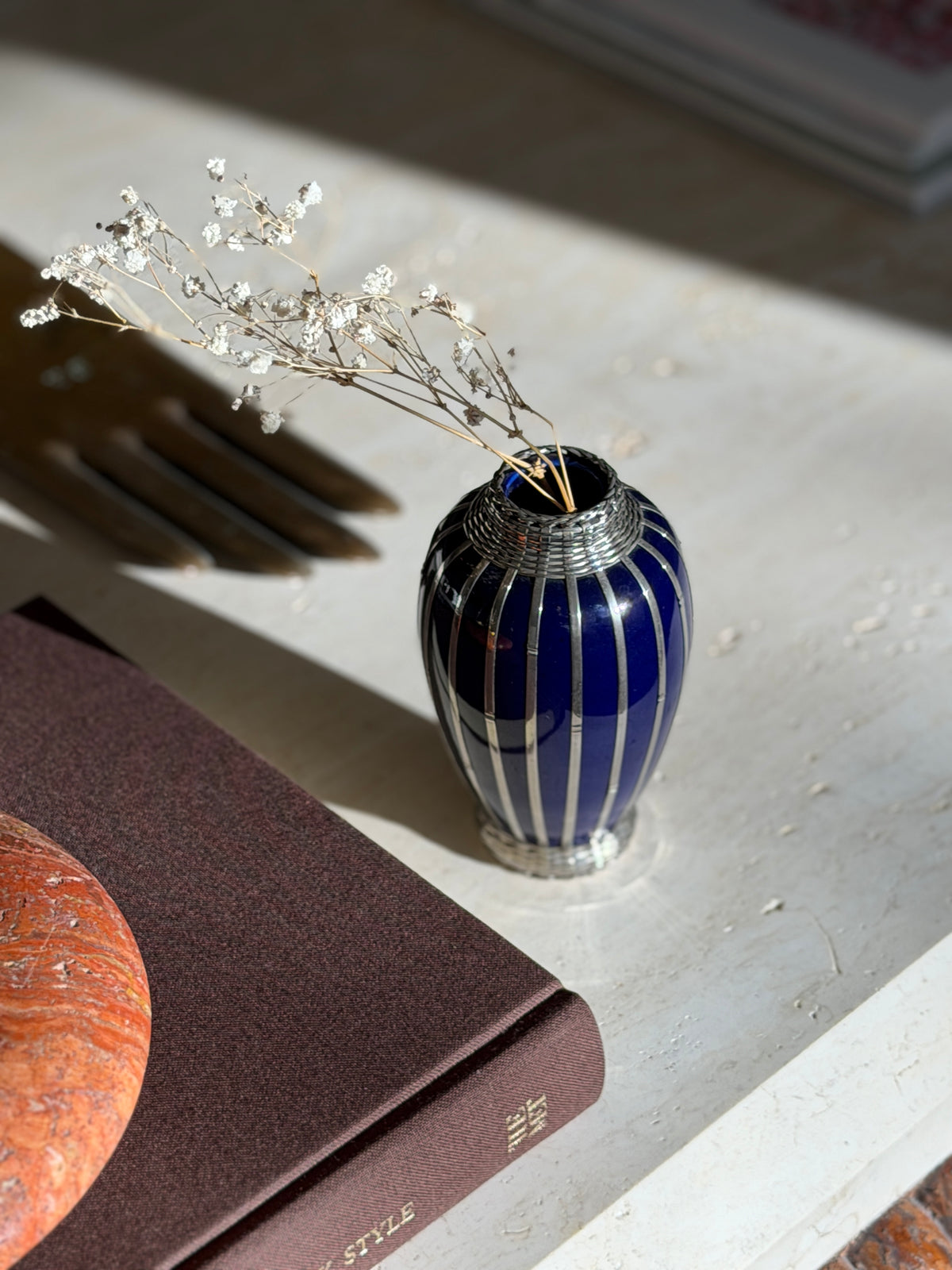 Overhead view of the silver overlay vase with dried flowers, the woven silver collar and ribbed body above a brown book
