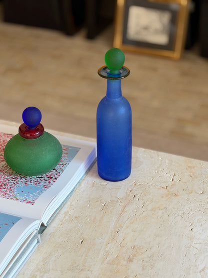 Tall cobalt blue scavo glass bottle with a frosted green ball stopper and flat amber-rimmed collar, standing at the edge of a travertine table