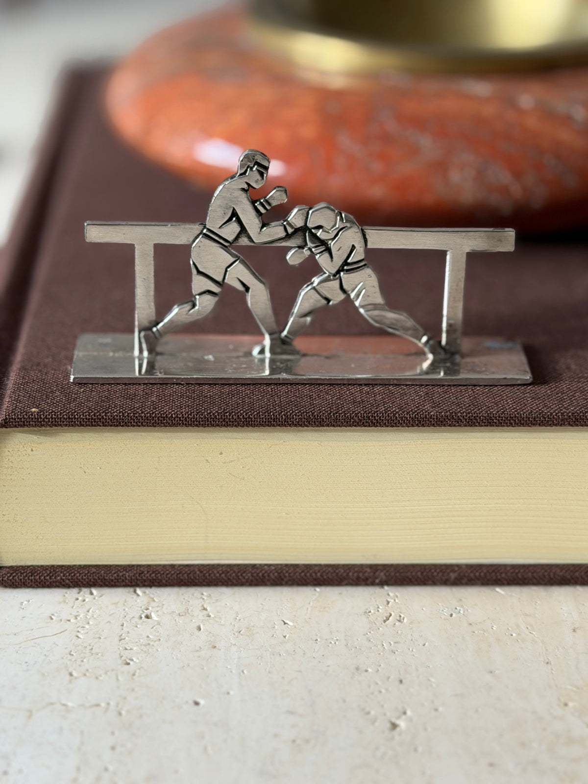Boxers knife rest on a brown clothbound book, one fighter throwing a punch as the other ducks behind the rail