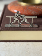 Boxers knife rest on a brown clothbound book, one fighter throwing a punch as the other ducks behind the rail