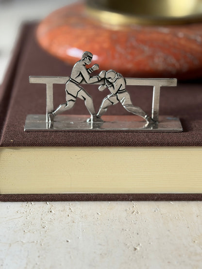 Boxers knife rest on a brown clothbound book, one fighter throwing a punch as the other ducks behind the rail