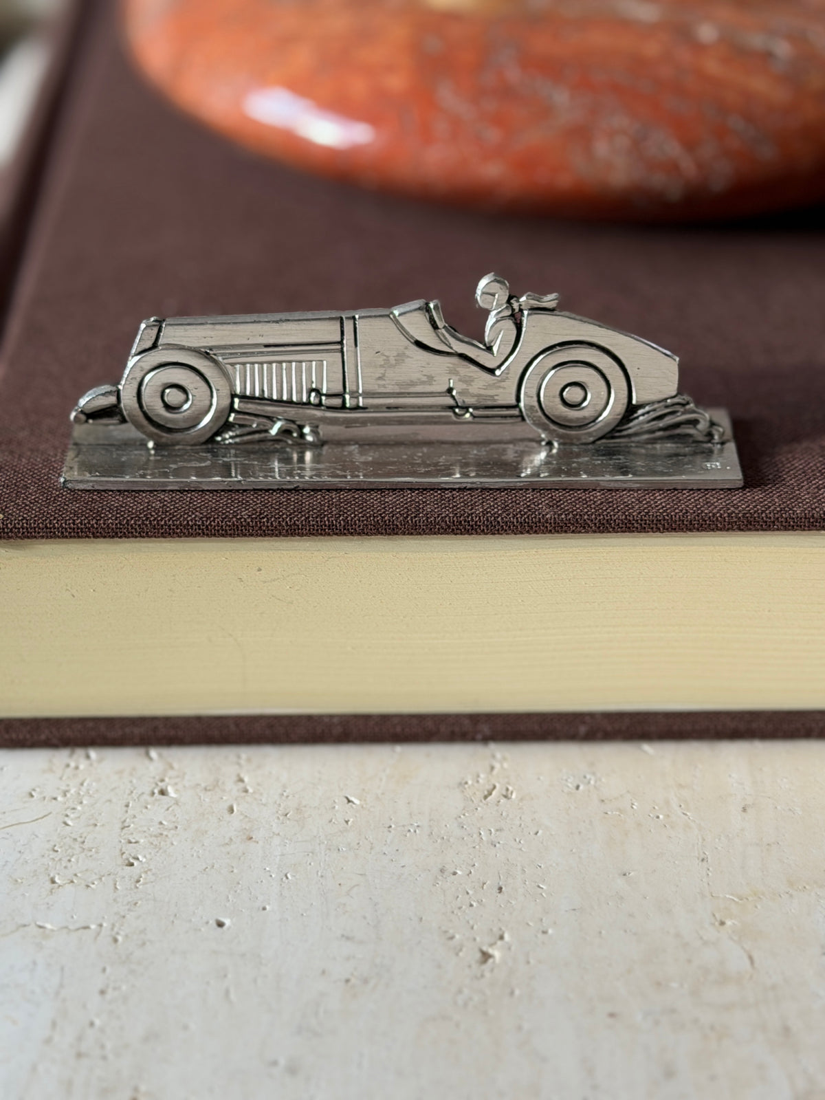 Race car knife rest on a brown book in profile, the louvered hood, round wheels and driver in relief, a red stone ashtray behind