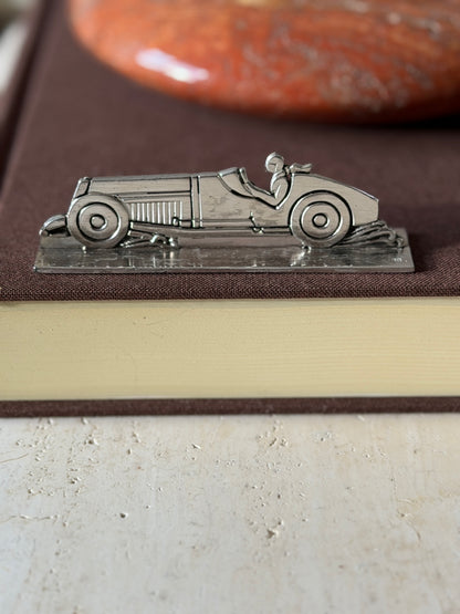 Race car knife rest on a brown book in profile, the louvered hood, round wheels and driver in relief, a red stone ashtray behind