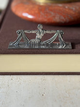 Front view of the draped dancer knife rest on a brown book, a small stamped mark at the corner of the base