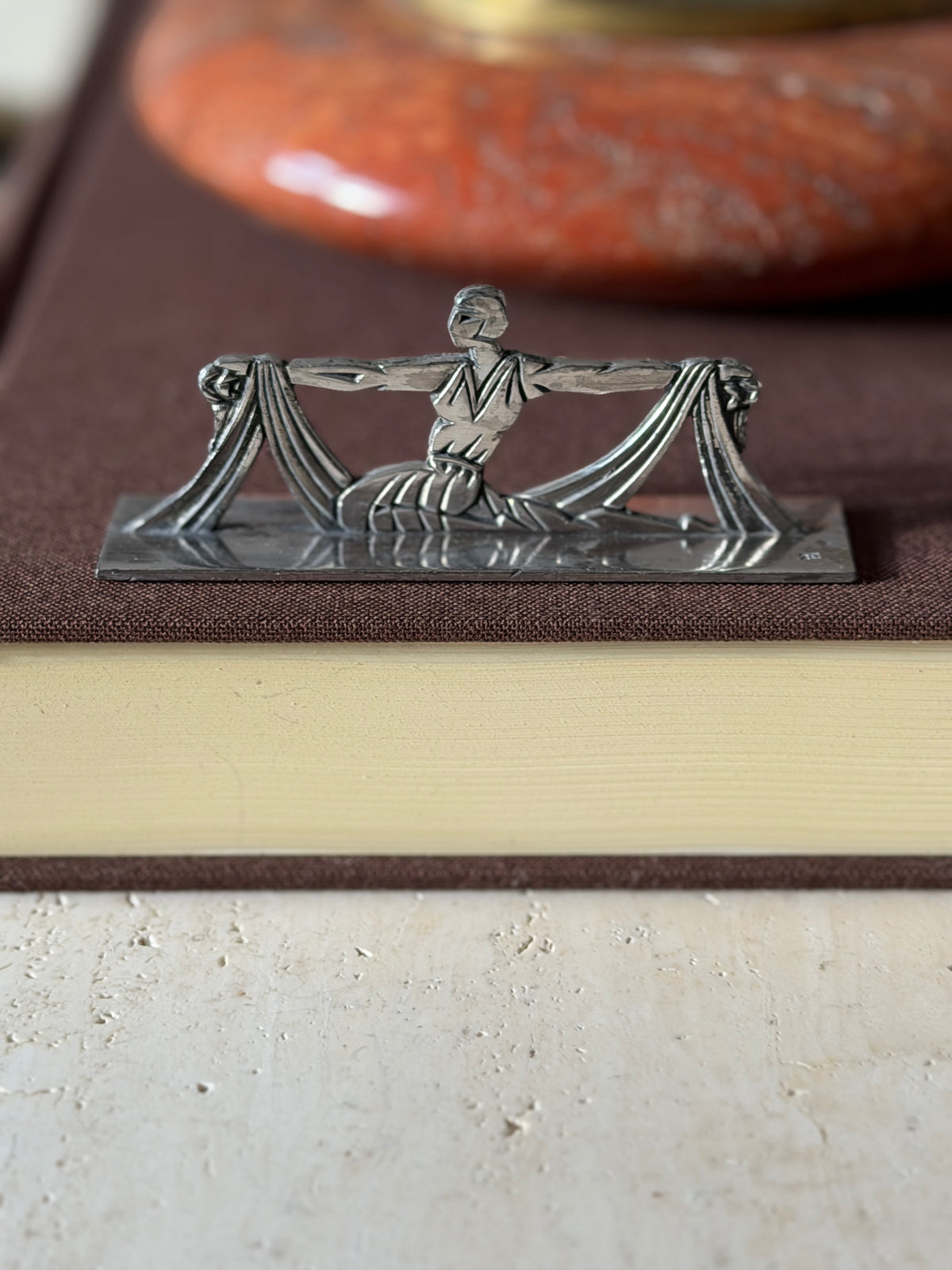Front view of the draped dancer knife rest on a brown book, a small stamped mark at the corner of the base