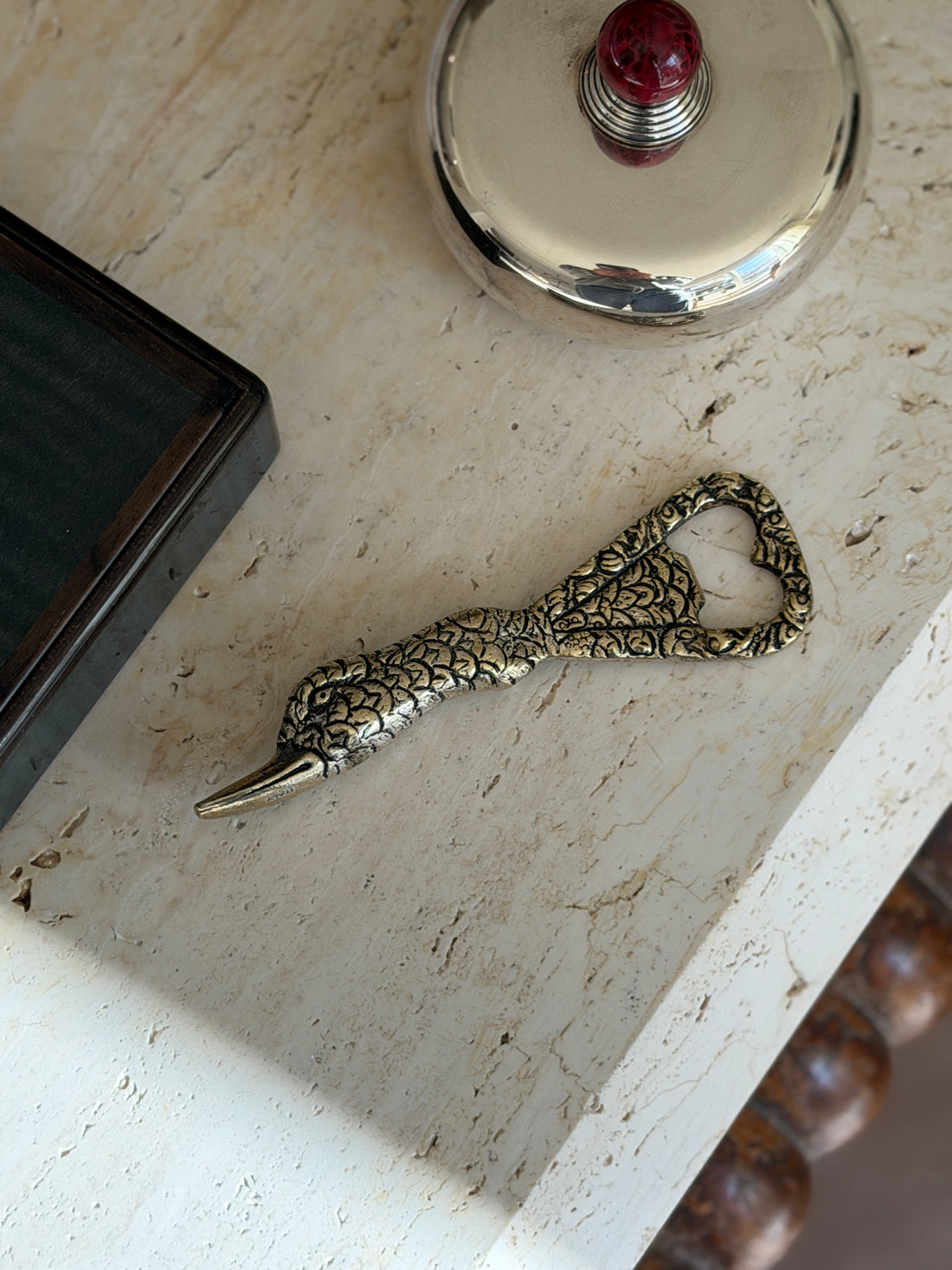 Gucci duck bottle opener in gilt brass on travertine, the feathered neck tapering to a long bill, near a dark wood box and a silver lidded box