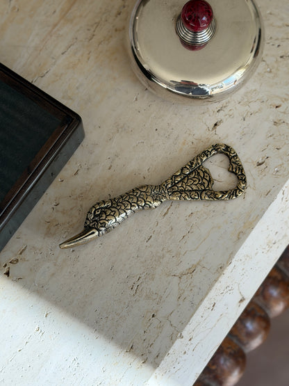 Gucci duck bottle opener in gilt brass on travertine, the feathered neck tapering to a long bill, near a dark wood box and a silver lidded box