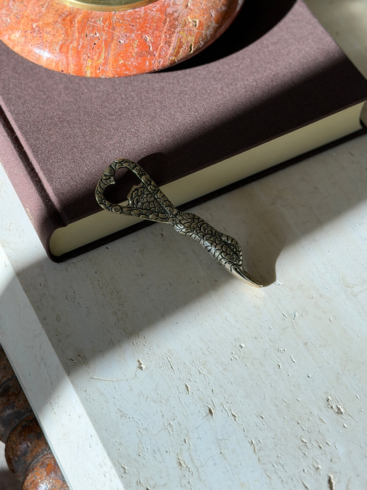 Gucci duck bottle opener leaning on a brown book beneath a red stone ashtray, its shadow cast across travertine