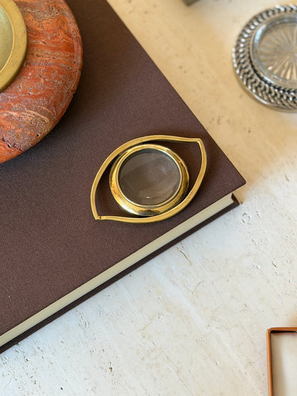 Eye-shaped gilt brass Hermes magnifier resting on a brown clothbound book beside a red stone ashtray and silver coaster