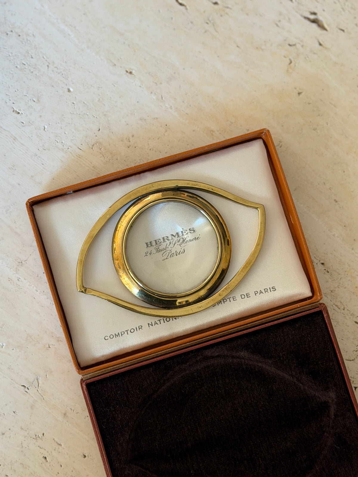 Hermes gilt magnifier laid on the satin lining of its box, the round lens enlarging the Hermes 24 Faubourg Saint-Honore Paris imprint