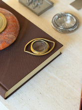 Angled view of the Cleopatras Eye magnifier on the corner of a brown book, the lens framed within the open almond-shaped brass outline