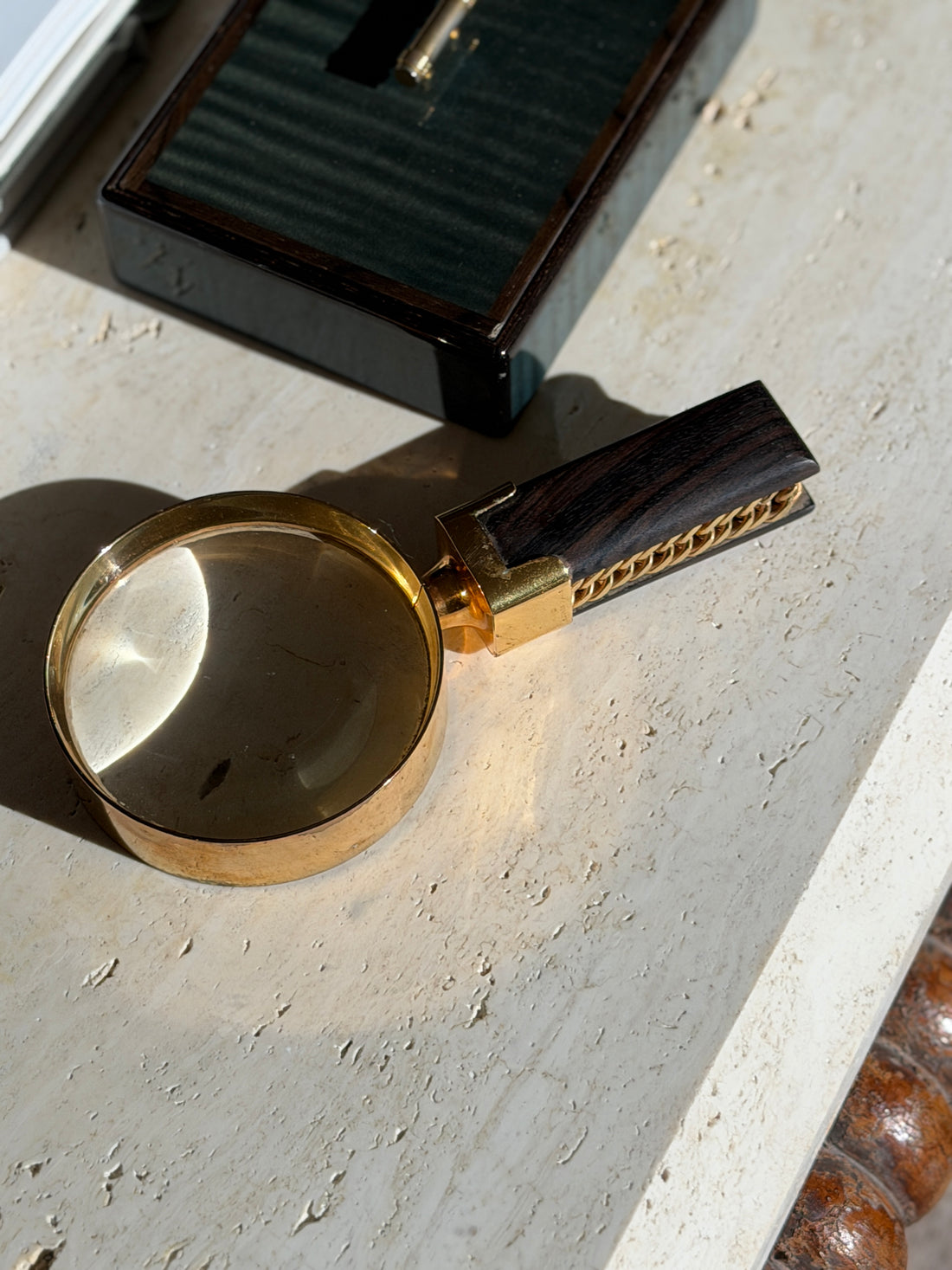Hermes magnifying glass with a gilt brass rim and a Macassar ebony handle inset with gold tone curb chain, on travertine near a lacquered box