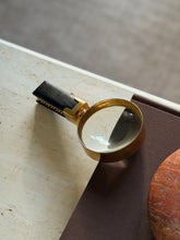 Gilt brass magnifier lying on the corner of a brown book, the domed lens catching window light and the chain trimmed ebony handle resting on travertine against a soft gray rug