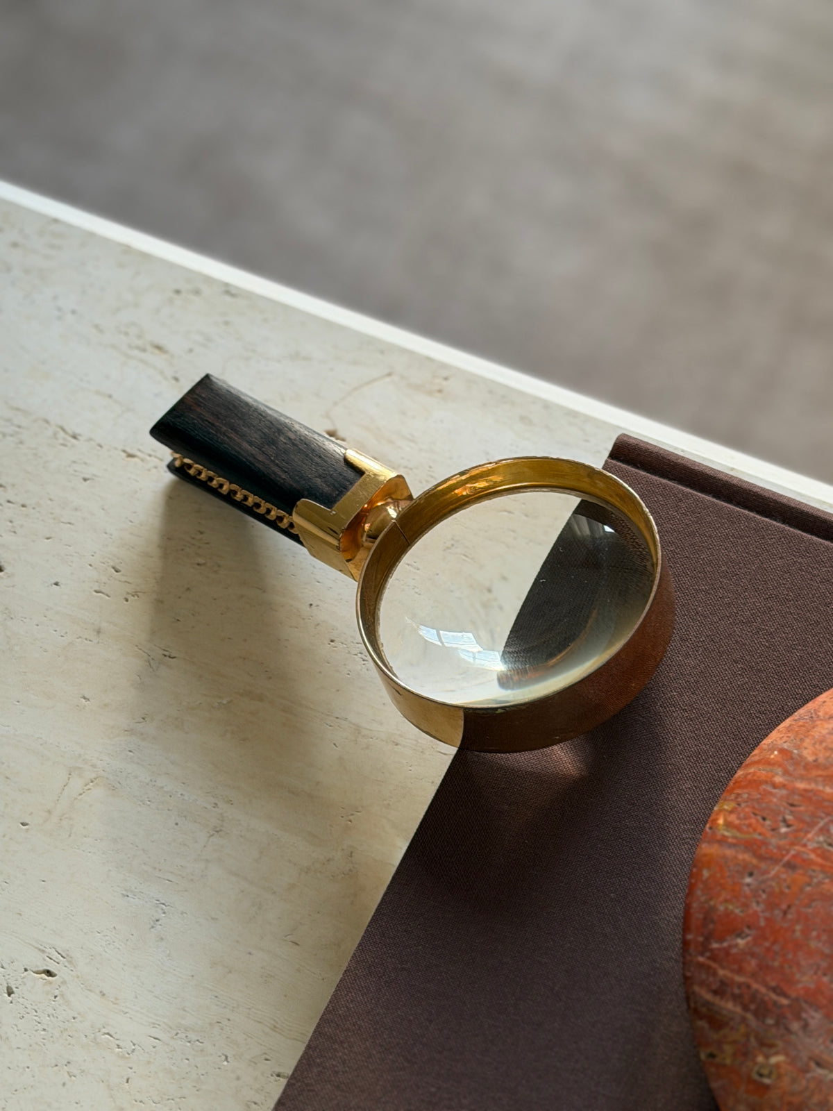 Gilt brass magnifier lying on the corner of a brown book, the domed lens catching window light and the chain trimmed ebony handle resting on travertine against a soft gray rug