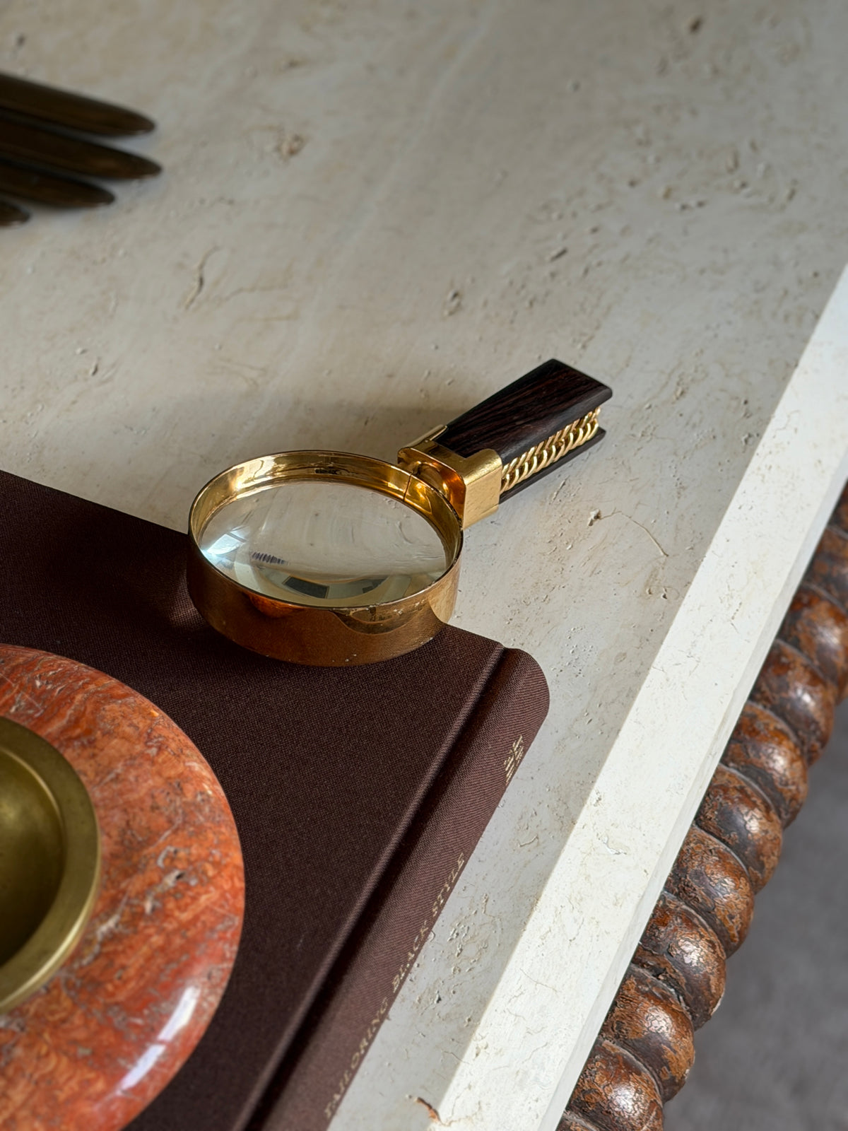 High angle view of the Macassar ebony and gilt brass magnifier on a brown book beside a red stone ashtray with brass well, the fingers of a brass glove mold at the top of the frame