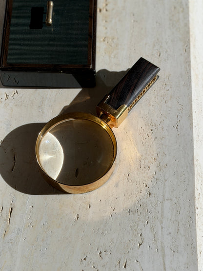 Overhead view of the Hermes magnifier on travertine, sunlight focused through the round lens beside a dark lacquered box